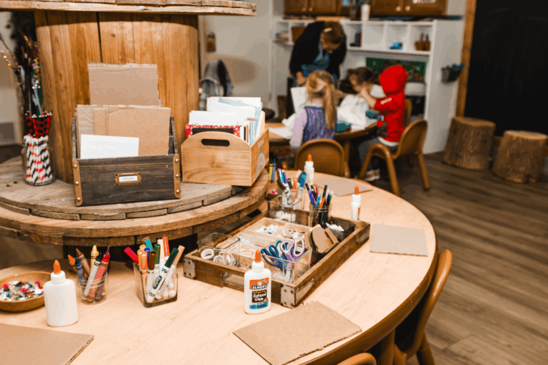 A classroom art station with markers, glue, scissors, and paper on tables; children and an adult work together at a table in the background.